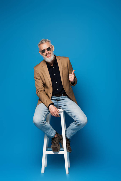 volle Länge des glücklichen Mannes mittleren Alters mit Sonnenbrille, der den Daumen nach oben zeigt, während er auf einem weißen Stuhl auf blau sitzt - Foto, Bild