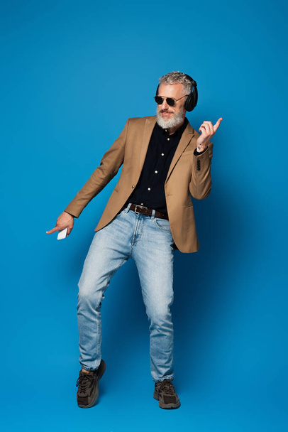 full length of smiling middle aged man in sunglasses holding smartphone and listening music in headphones on blue - Foto, Imagem