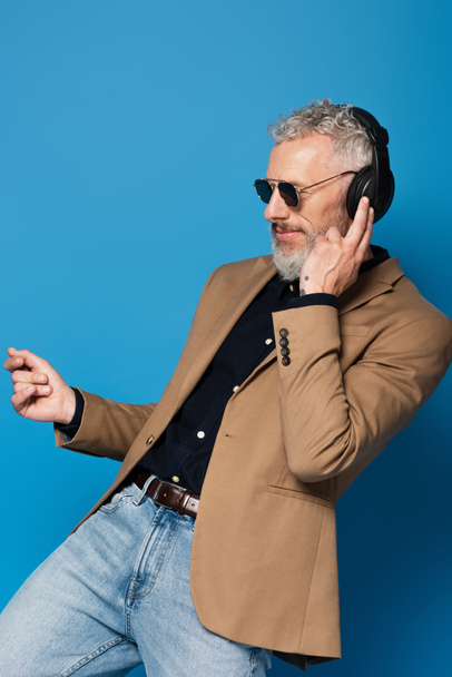 smiling middle aged man in sunglasses listening music in headphones while dancing isolated on blue - Foto, Imagem