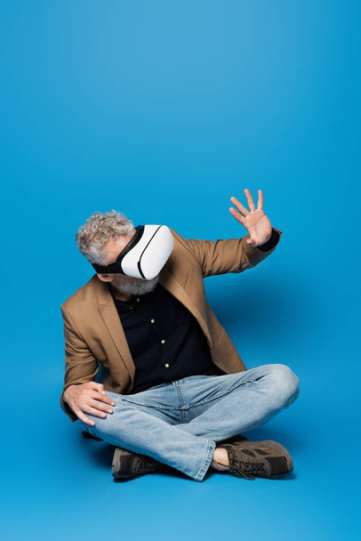 full length of middle aged man in vr headset sitting with crossed legs and gesturing on blue  - Photo, Image