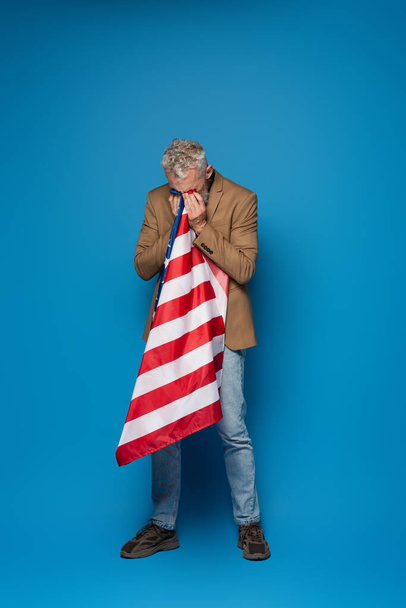 full length of patriotic middle aged man crying and covering face with american flag on blue  - Photo, image
