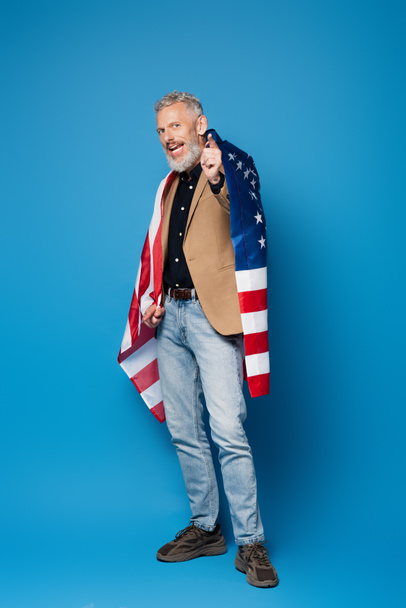 full length of bearded middle aged man standing with american flag and pointing at camera on blue  - Zdjęcie, obraz