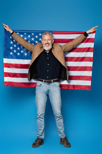 longitud completa de hombre de mediana edad complacido de pie con bandera americana en azul  - Foto, imagen