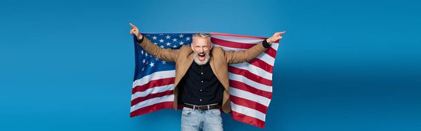 excited middle aged man standing with american flag on blue, banner - Foto, imagen