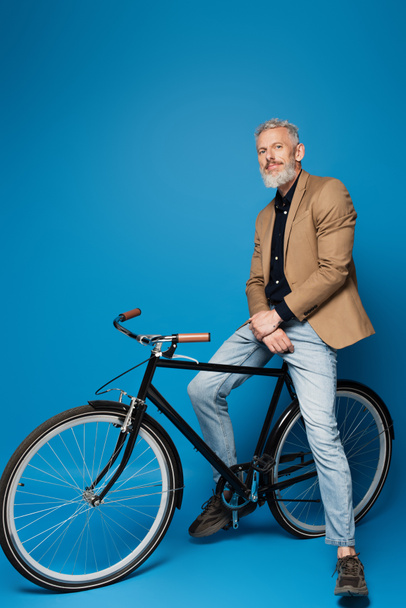 full length of pleased middle aged man sitting on bicycle on blue  - Фото, зображення