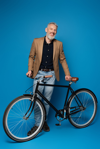 pełna długość zadowolony mężczyzna w średnim wieku stojący w pobliżu roweru na niebiesko  - Zdjęcie, obraz