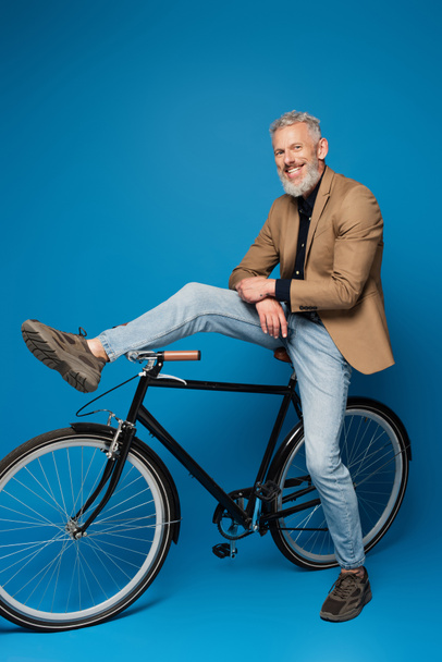 full length of cheerful middle aged man sitting on bicycle on blue  - Fotografie, Obrázek