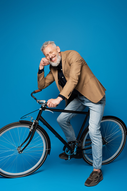 full length of pleased middle aged man sitting on bicycle and smiling on blue  - 写真・画像