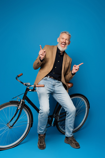 full length of joyful middle aged man sitting on bicycle and pointing with fingers on blue  - Foto, imagen