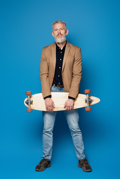 full length of bearded middle aged man with grey hair standing with longboard on blue  - Foto, Bild