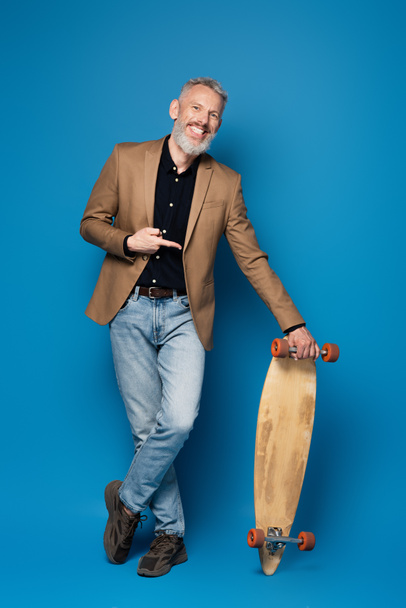 full length of pleased middle aged man pointing at longboard on blue  - Fotografie, Obrázek