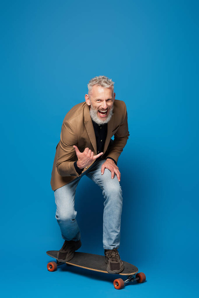 longitud completa de hombre de mediana edad excitado montando en longboard y gestos en azul  - Foto, Imagen