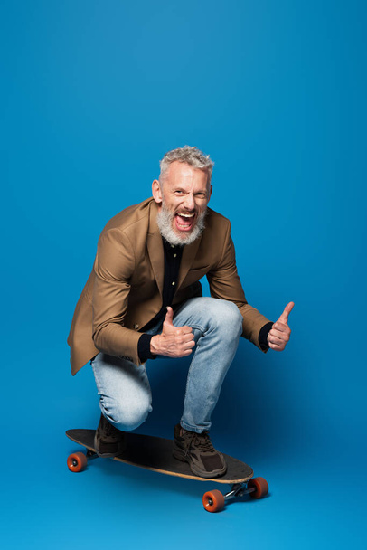 full length of pleased middle aged man riding on longboard and showing thumbs up on blue  - Photo, Image
