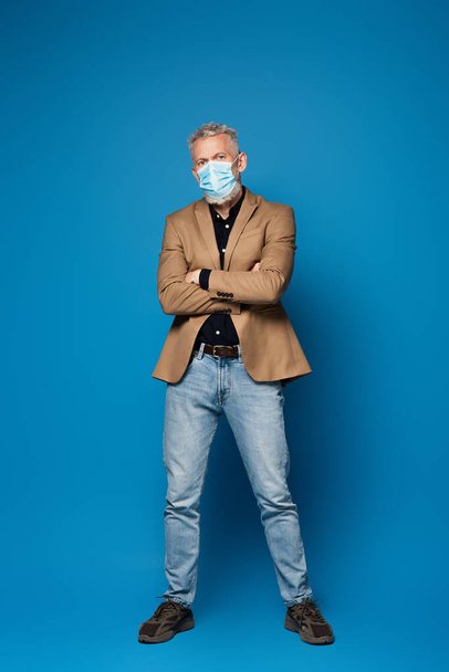 plná délka muže středního věku v ochranné lékařské masce stojící se zkříženýma rukama na modré  - Fotografie, Obrázek