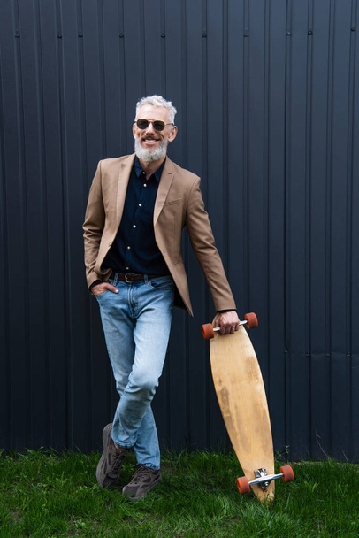 full length of pleased middle aged man in sunglasses holding longboard outside  - Фото, зображення