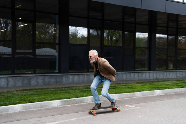 full length of happy middle aged man in sunglasses riding longboard outside  - Photo, Image