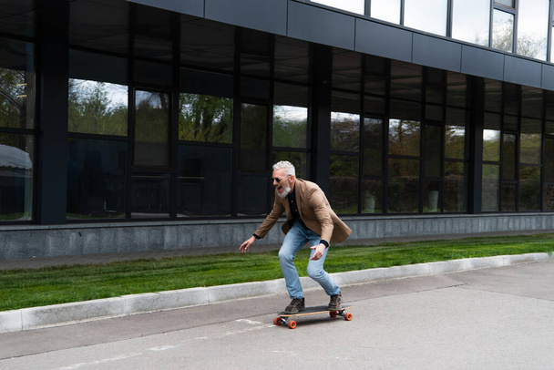 full length of positive middle aged man in sunglasses riding longboard outside  - Photo, Image