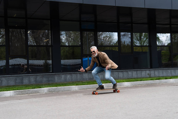 full length of happy middle aged man in sunglasses riding longboard and gesturing outside  - 写真・画像