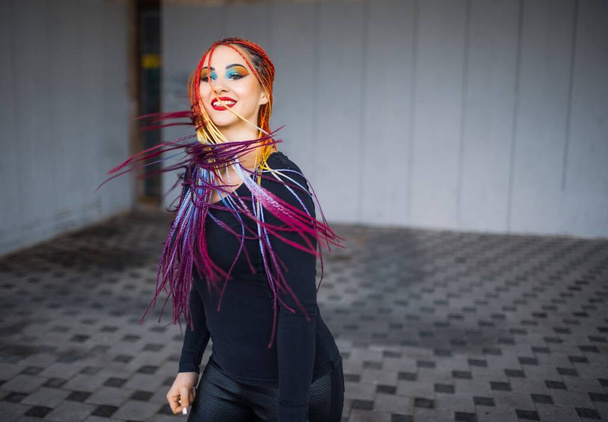 The most beautiful girl in a black elegant dress with bright makeup and multi-colored artificial braids. Beautifully circling and waving long braids against the backdrop of an old building. - Фото, изображение