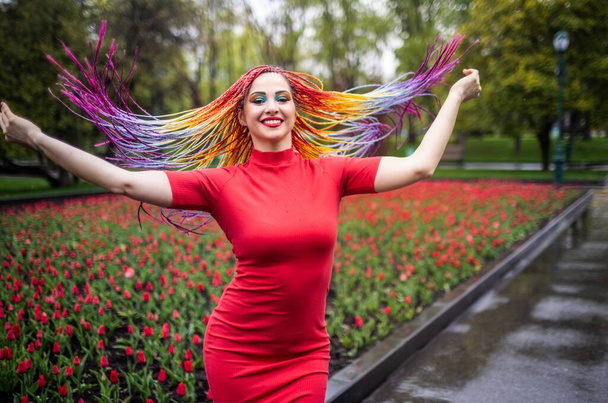 A gentle girl with expressive shiny makeup and multi-colored long braids in red dress. Swirls in spring park against the backdrop of a blooming flower bed with tulips and enjoy the arrival of spring. - Foto, immagini