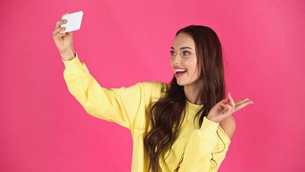 smiling young adult woman taking selfie on cellphone with peace gesture isolated on pink - Photo, image