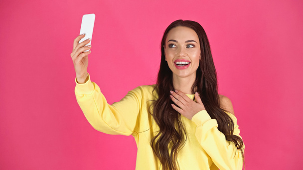 smiling young adult woman taking selfie on cellphone and holding hand on chest isolated on pink - Φωτογραφία, εικόνα