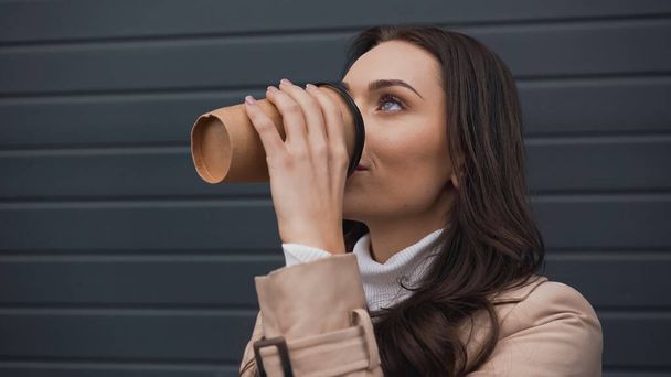 young adult woman in trench coat drinking coffee from paper cup on grey textured background - Foto, immagini