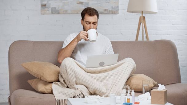 sick man sitting on sofa with laptop and drinking tea near coffee table with pills in bottles  - Valokuva, kuva