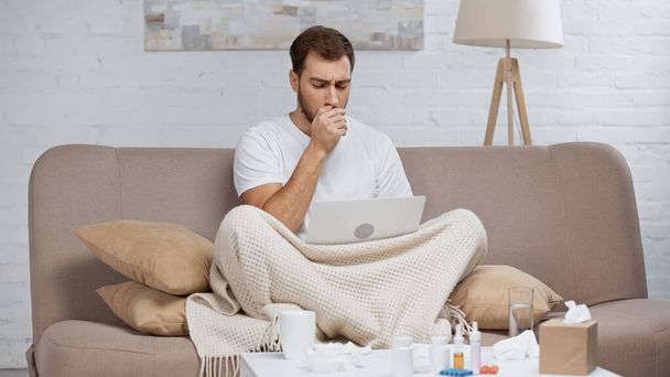 sick freelancer sitting on sofa and using laptop while coughing near coffee table with drinks and bottles  - 写真・画像