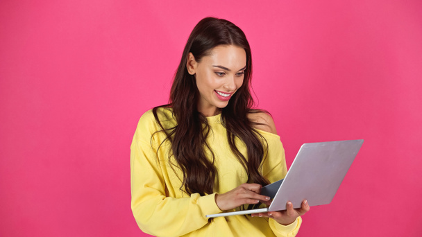 smiling young adult woman typing on laptop and holding credit card isolated on pink - Foto, afbeelding
