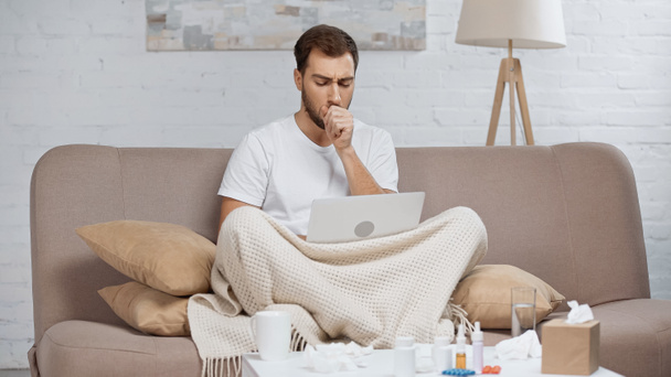 sick freelancer sitting on sofa and using laptop while coughing near coffee table with drinks and bottles  - 写真・画像