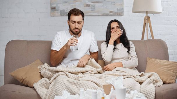 sick man holding glass of water near diseased woman sneezing in living room  - Fotó, kép