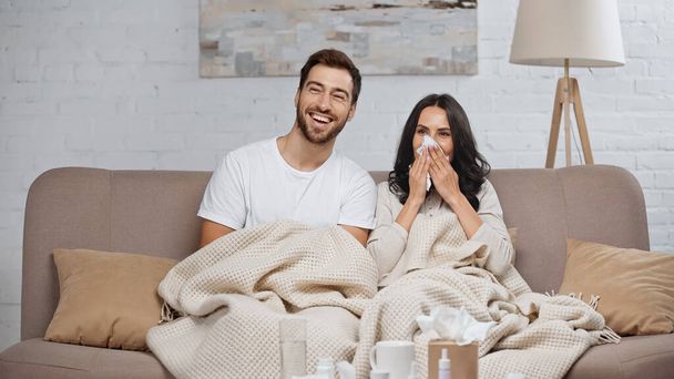 bearded man smiling near sick girlfriend sneezing in tissue  - Photo, image