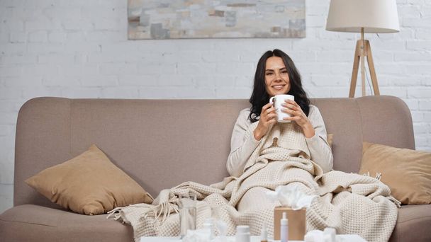 sick brunette woman holding cup of tea and smiling while sitting on couch - Foto, imagen