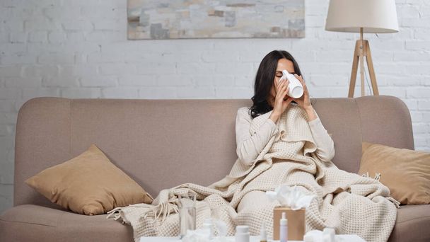 sick brunette woman holding cup and drinking tea while sitting on couch - Fotó, kép