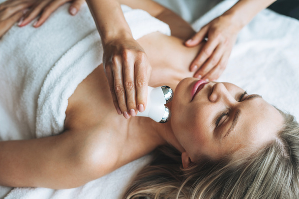 Giovane donna bionda gode di massaggio facciale con microcorrente massaggiatore facciale nella spa, concedetevi. Cura del corpo professionale - Foto, immagini