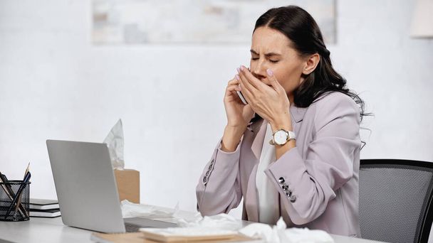 brunette businesswoman with allergy sneezing while talking on smartphone near laptop on desk - 写真・画像