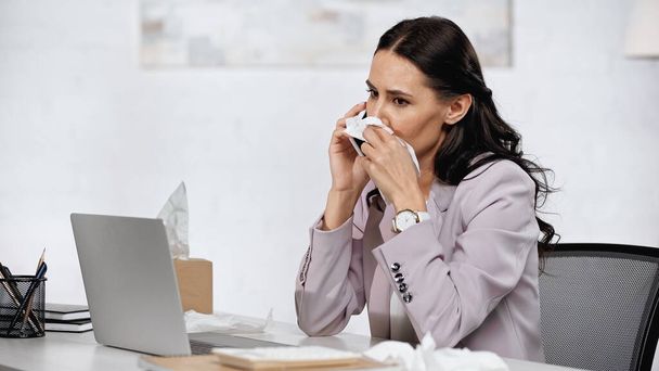brunette businesswoman with allergy sneezing in napkin while talking on cellphone near laptop on desk - 写真・画像