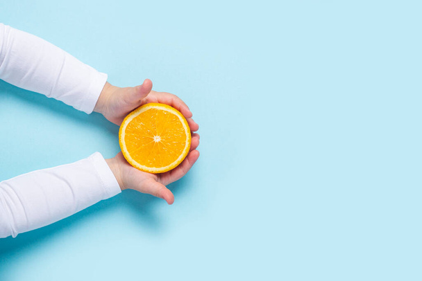 children's hands hold a cut half of an orange on a blue background. Top view, flat lay. - 写真・画像