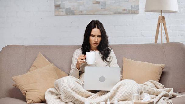 sick woman sitting with laptop and holding cup of tea  - Fotoğraf, Görsel