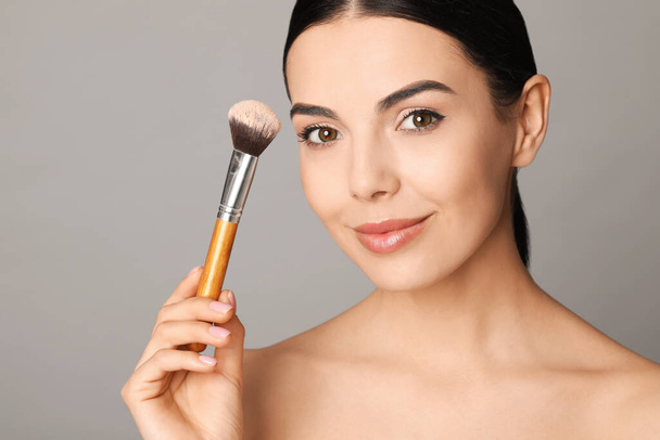 Beautiful young woman applying face powder with brush on grey background, closeup - Photo, image