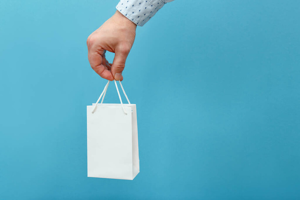A hand holds a gift in a white bag on a blue background. - Foto, afbeelding