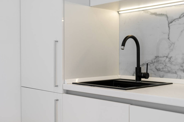 Fully furnished modern white kitchen with black sink in minimalistic theme built in countertop, lit with soft light above table - Photo, Image