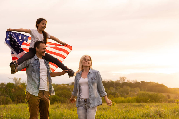 Młodzi rodzice z córką trzymają amerykańską flagę na wsi o zachodzie słońca. Obchody Dnia Niepodległości - Zdjęcie, obraz