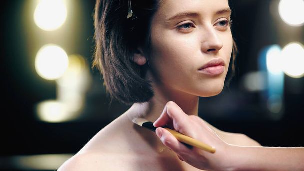 maquillador aplicando fundación sobre clavículas de modelo morena con pincel cosmético - Foto, Imagen