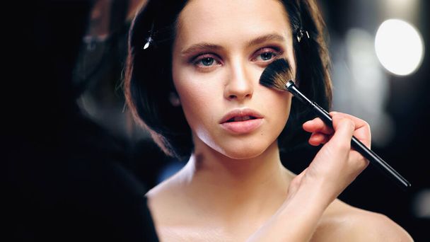 maquillador aplicando polvo facial en la cara del modelo con hombros desnudos - Foto, imagen