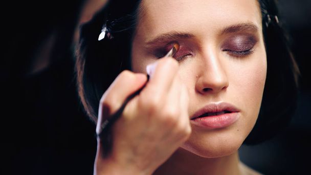makeup artist applying eye shadow with cosmetic brush on model with closed eyes - Fotó, kép
