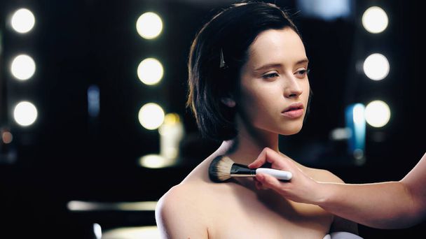 makeup artist applying powder on body of brunette model with bare shoulders - Foto, Imagen