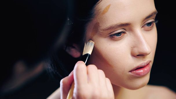 Bulanık makyaj sanatçısı kozmetik fırçayla genç modelin yüz hatlarını çiziyor. - Fotoğraf, Görsel