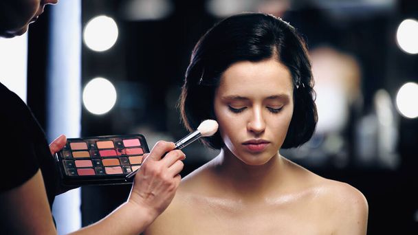 makeup artist holding palette and applying highlighter on skin of model with cosmetic brush - Photo, Image
